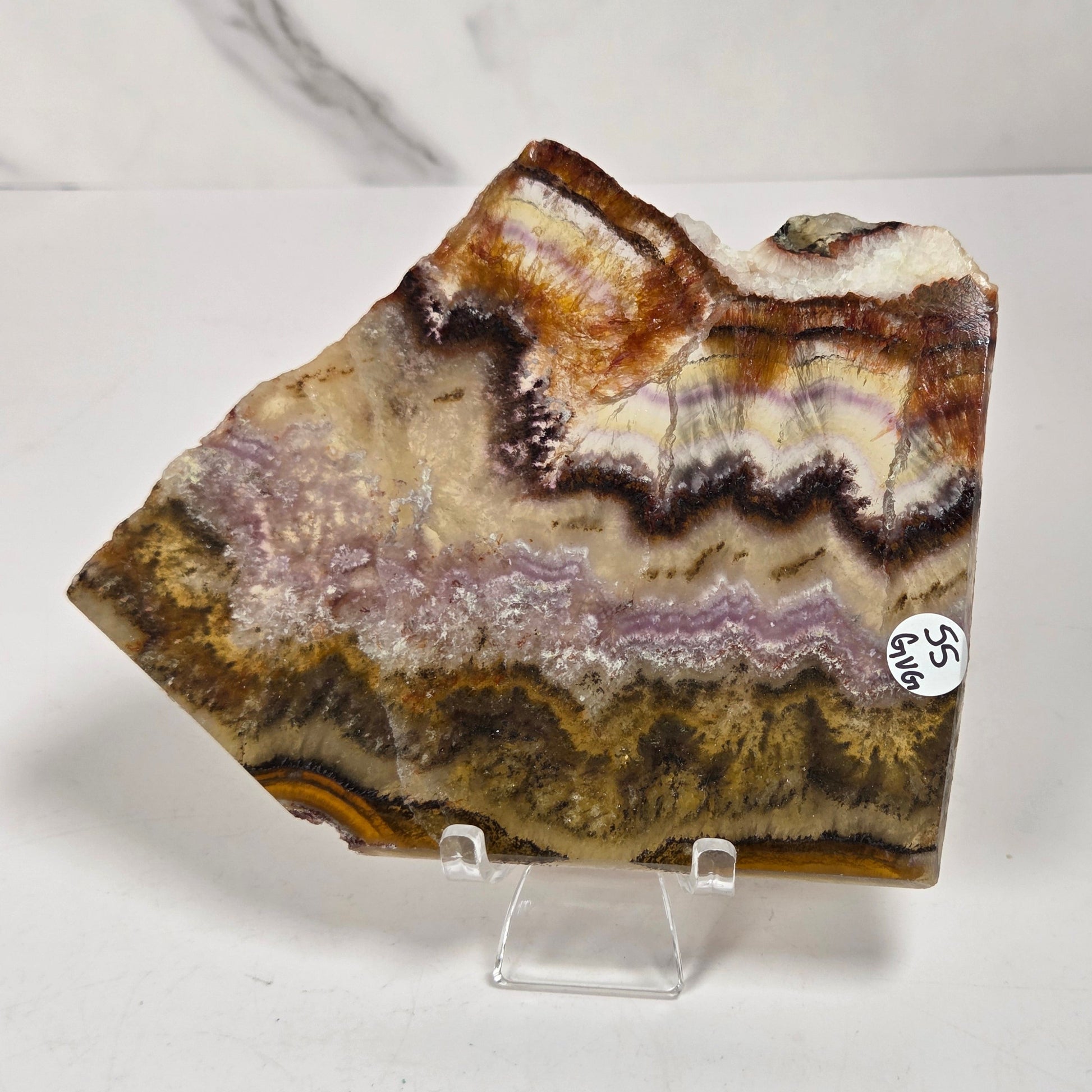 A raw agate crystal specimen with layered bands of brown, beige, purple, and gold, displayed on a clear acrylic stand against a white marble-patterned background.