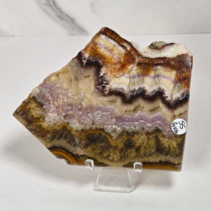 A raw agate crystal specimen with layered bands of brown, beige, purple, and gold, displayed on a clear acrylic stand against a white marble-patterned background.