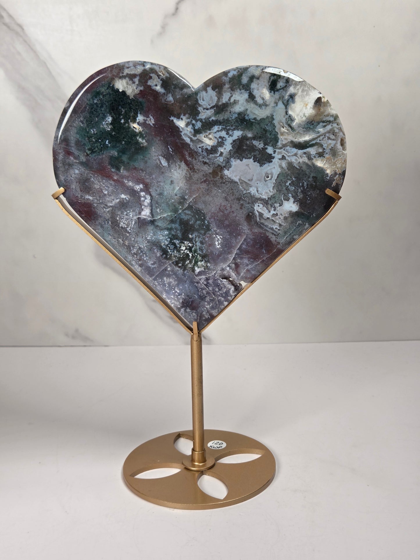 Heart-shaped polished stone sculpture on a gold metal stand, featuring a marbled pattern of gray, blue, and purple tones, set against a light marble-patterned background.