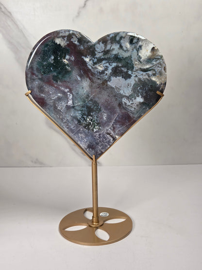 Heart-shaped polished stone sculpture on a gold metal stand, featuring a marbled pattern of gray, blue, and purple tones, set against a light marble-patterned background.