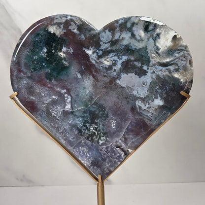 Heart-shaped polished stone plaque with marbled gray, blue, green, and purple tones, displayed on a gold-toned stand against a light marble-patterned background.