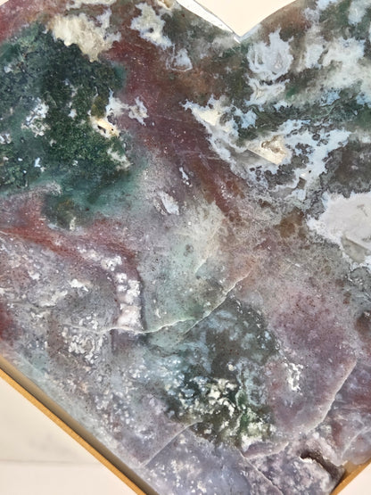 A close-up of a polished stone or mineral slab with marbled patterns in green, gray, purple, and reddish-brown, filling most of the frame.