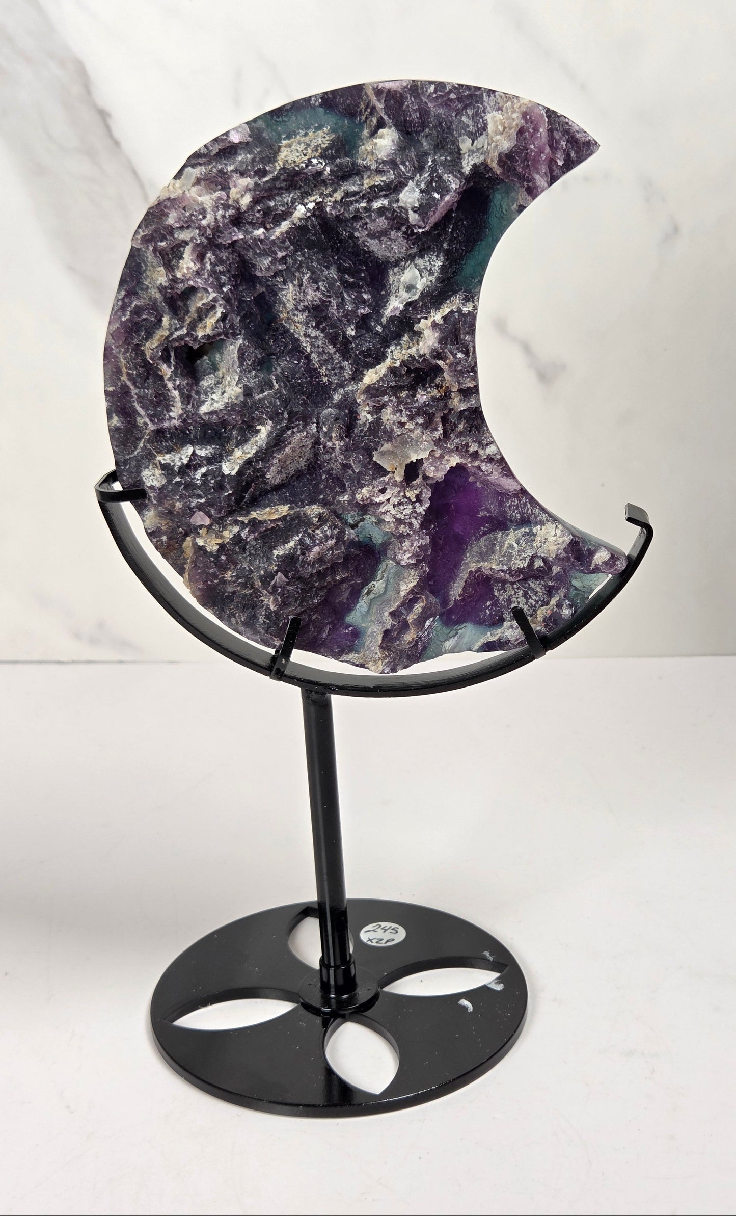 A crescent moon-shaped polished stone plaque with natural purple, gray, and white marbling, displayed on a black metal stand against a light marble-patterned background.