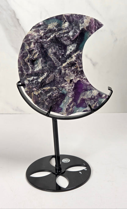 A crescent moon-shaped polished stone plaque with natural purple, gray, and white marbling, displayed on a black metal stand against a light marble-patterned background.