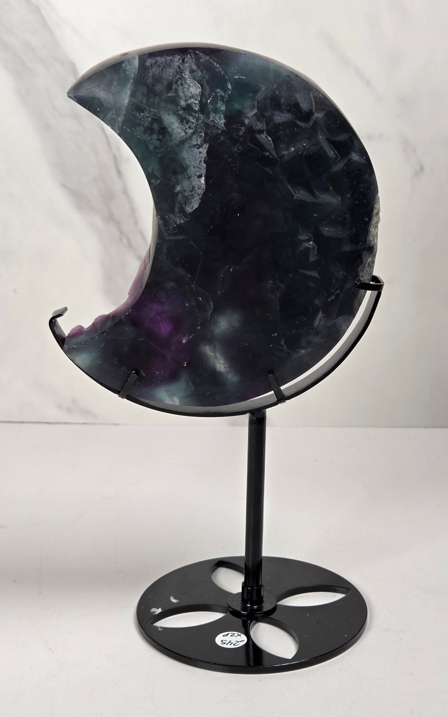 A crescent moon-shaped polished stone sculpture with a black metal stand, featuring a marbled pattern of black, gray, and purple, set against a light gray marble-patterned background.