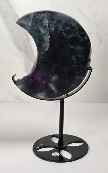 A crescent moon-shaped polished stone sculpture with a black metal stand, featuring a marbled pattern of black, gray, and purple, set against a light gray marble-patterned background.