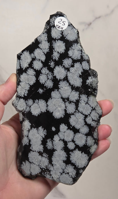 A raw mineral specimen of black obsidian with white to light-gray dendritic crystalline patterns, displayed on a clear acrylic stand against a white marble background.