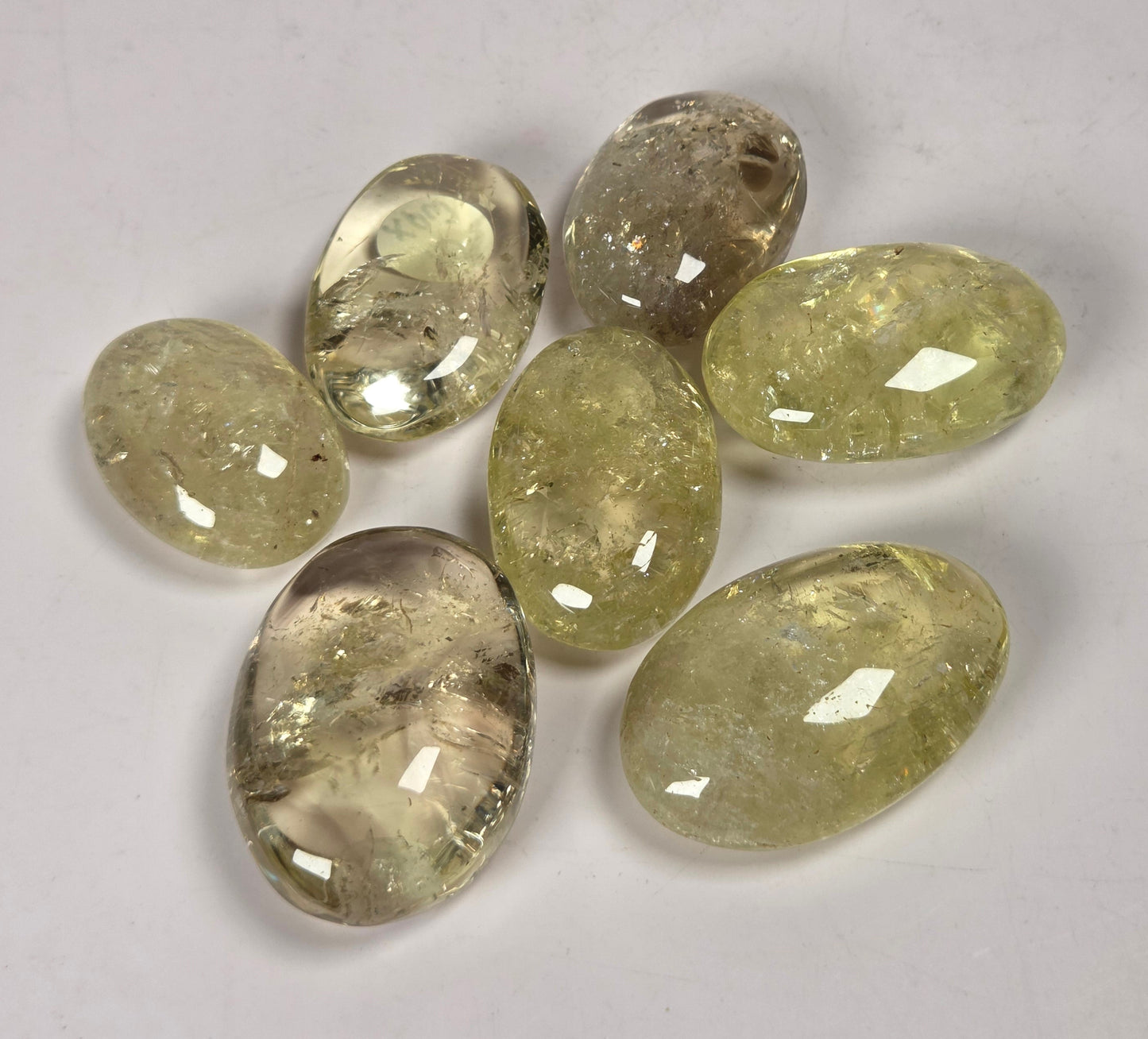 Seven faceted oval gemstones in a light yellow-green hue with internal inclusions, arranged on a plain light gray background.