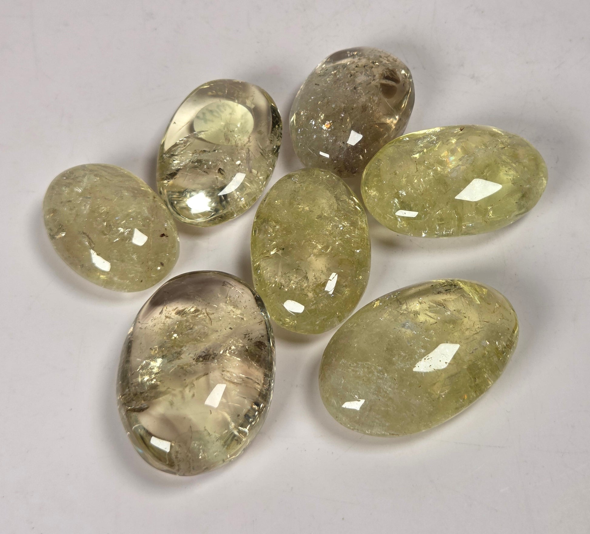 Seven faceted oval gemstones in a light yellow-green hue with internal inclusions, arranged on a plain light gray background.