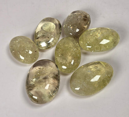 Seven faceted oval gemstones in a light yellow-green hue with internal inclusions, arranged on a plain light gray background.