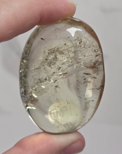 A hand holding a polished oval clear gemstone with internal inclusions and a metallic gold-toned center, photographed against a light gray marble-like background.