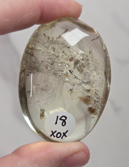 A hand holds a polished, oval-shaped clear stone or resin piece containing natural inclusions and fibers. A small white sticker with the handwritten text '18 xox' is attached near the bottom of the specimen.