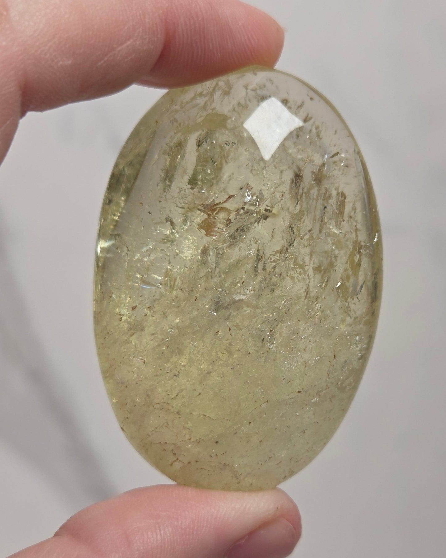 A hand holding a polished oval-shaped clear crystal stone with internal inclusions, set against a light gray marble background.