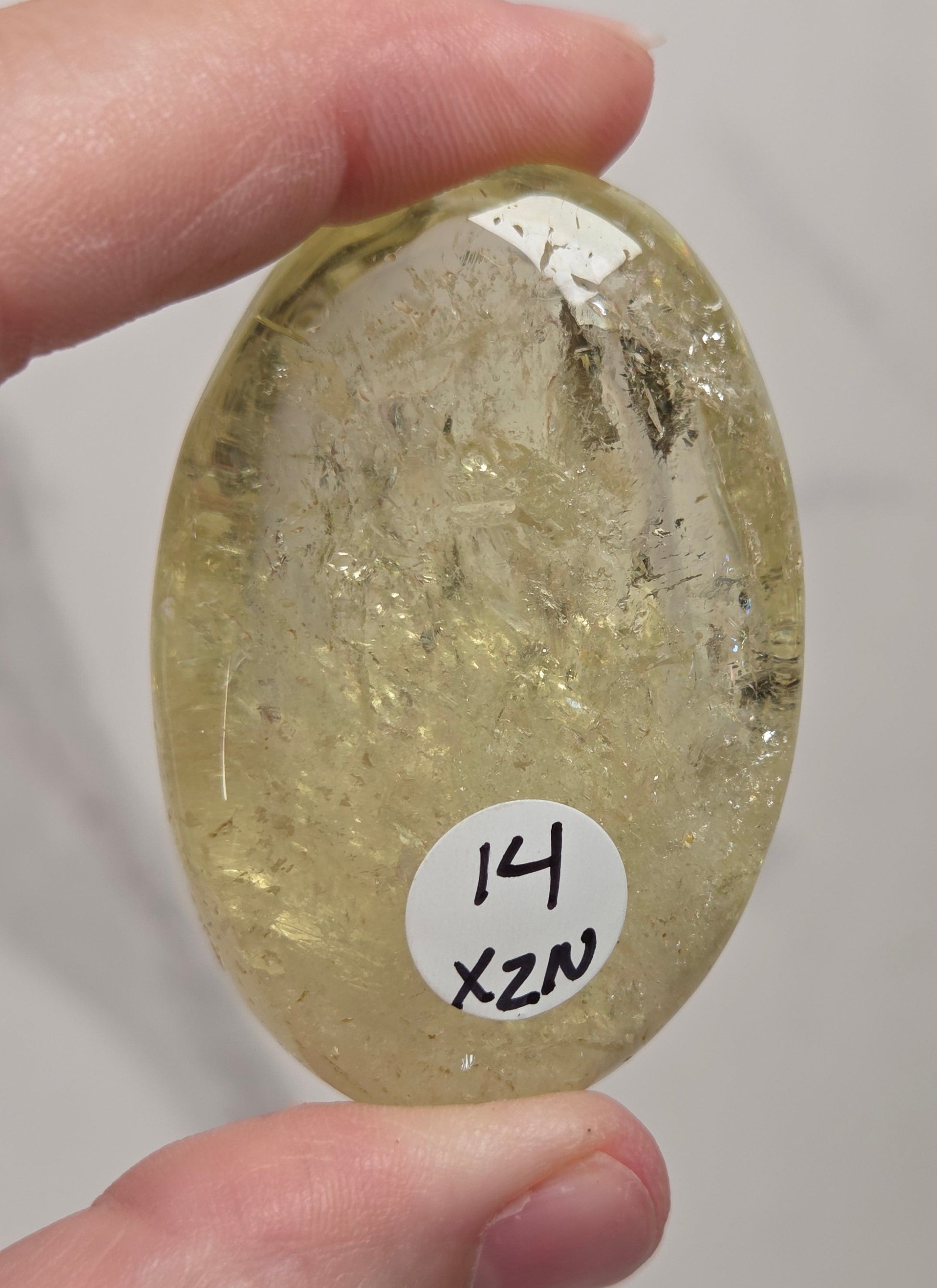 A hand holds a polished oval yellow stone with internal inclusions and a small white label reading '14 X2N' against a light neutral background.