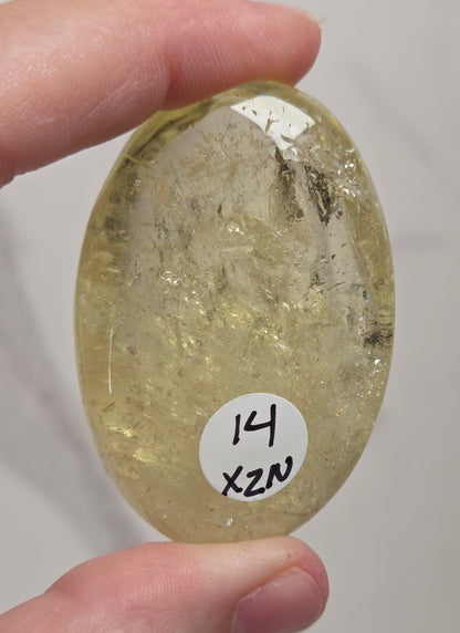 A hand holds a polished oval yellow stone with internal inclusions and a small white label reading '14 X2N' against a light neutral background.