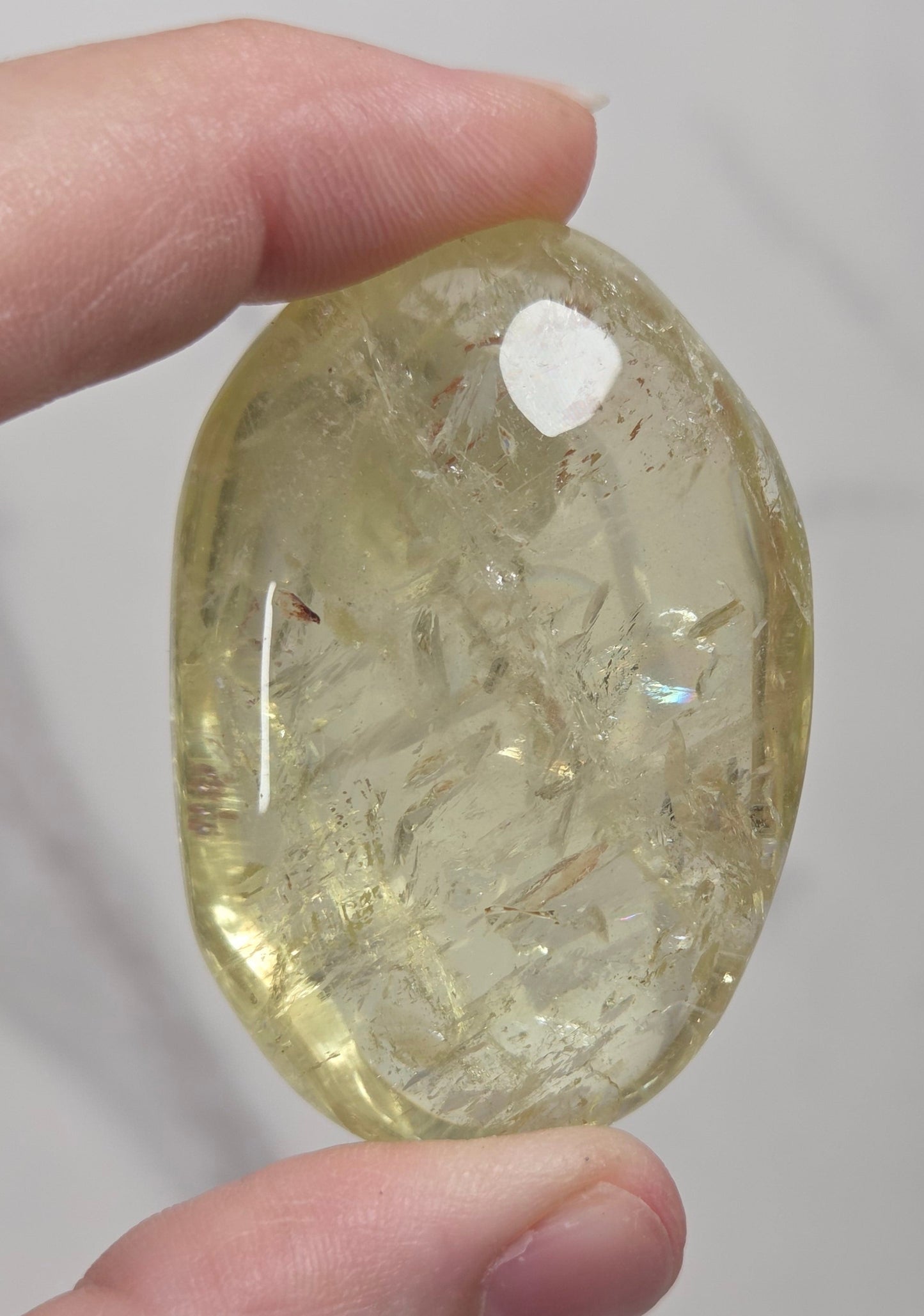 A hand holding a polished, oval-shaped yellow crystal stone with visible internal inclusions against a light gray marble background.