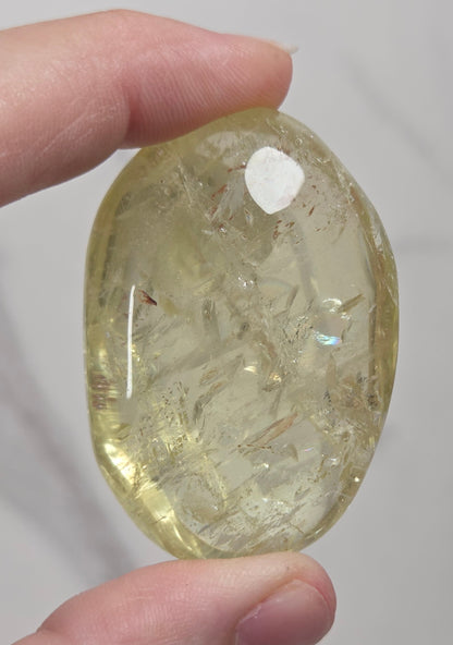 A hand holding a polished, oval-shaped yellow crystal stone with visible internal inclusions against a light gray marble background.