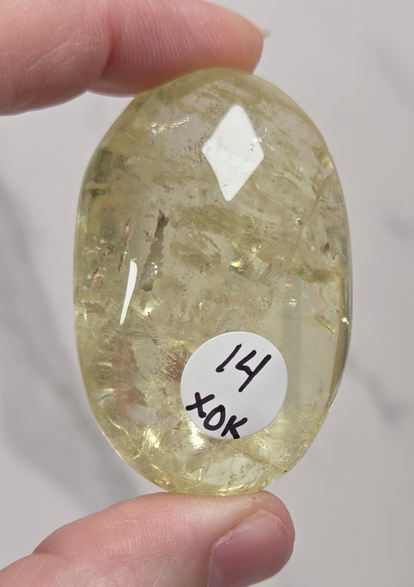 A hand holding a faceted, oval-shaped pale yellow gemstone with a small white label that reads '14 XOK' on a light gray marble background.