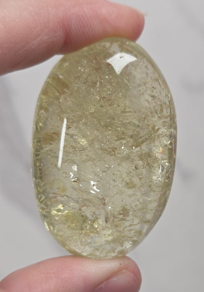 A hand holding a polished oval cabochon gemstone with a milky, translucent golden-yellow hue and internal inclusions, set against a light gray background.