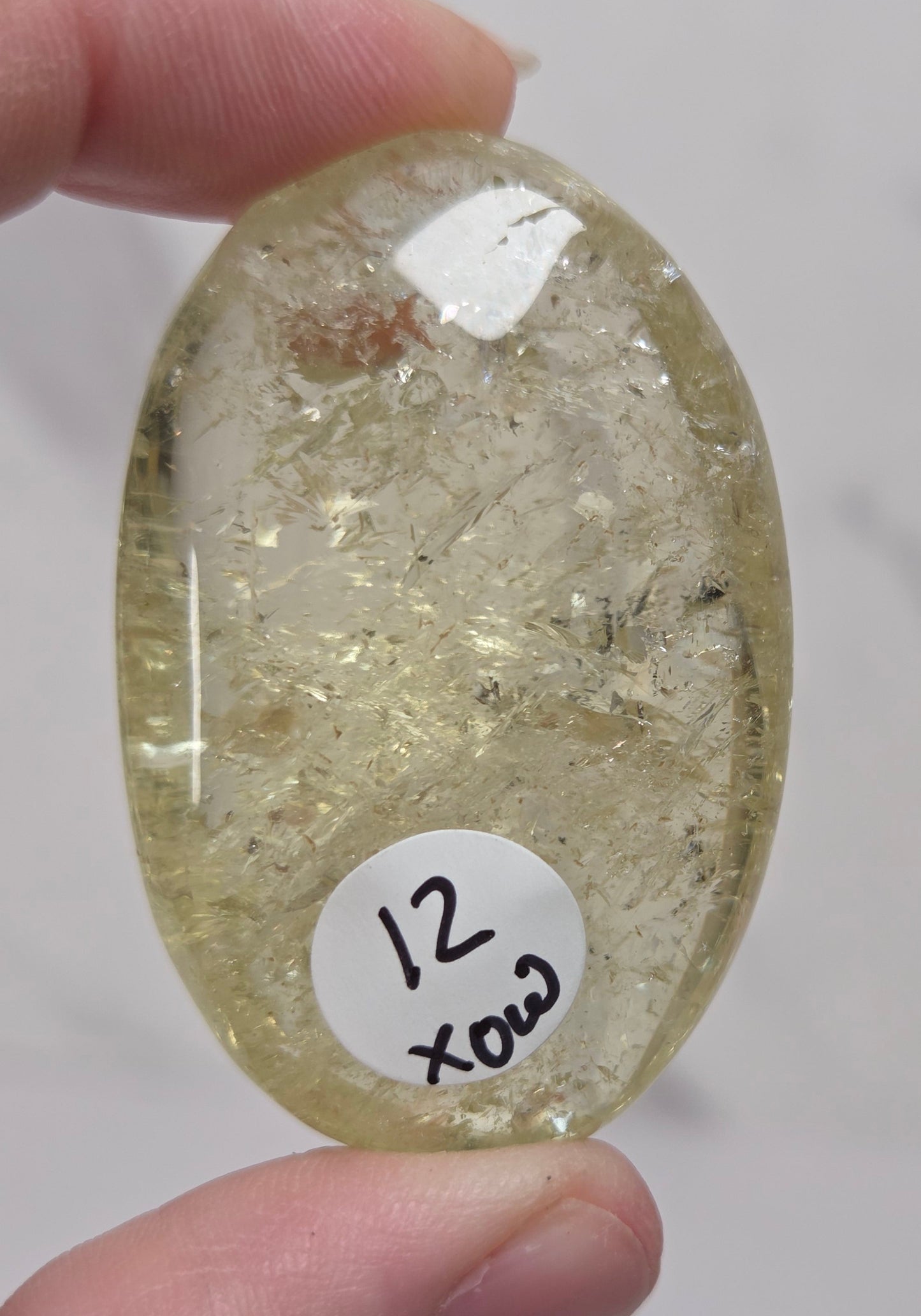 A hand holds a polished oval gemstone cabochon with a pale yellow-green hue and internal inclusions, featuring a white sticker with handwritten text '12 xod'.