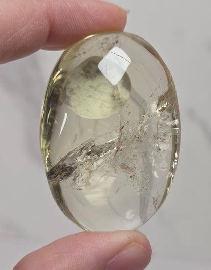 A hand holding a polished oval clear stone with internal inclusions and a warm yellow tint, photographed against a light gray marble-like background.