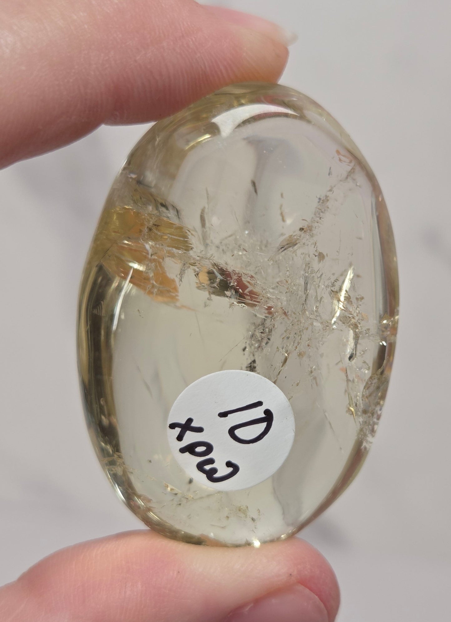A hand holds a small, polished oval stone or cabochon with a white sticker reading '10 xpcs' on it. The stone is transparent with light yellowish-brown inclusions.