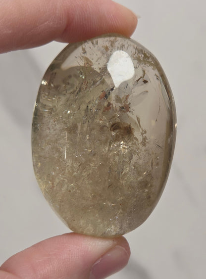 A hand holding a polished oval gemstone with internal inclusions and a white flash reflection, set against a light gray marble background.