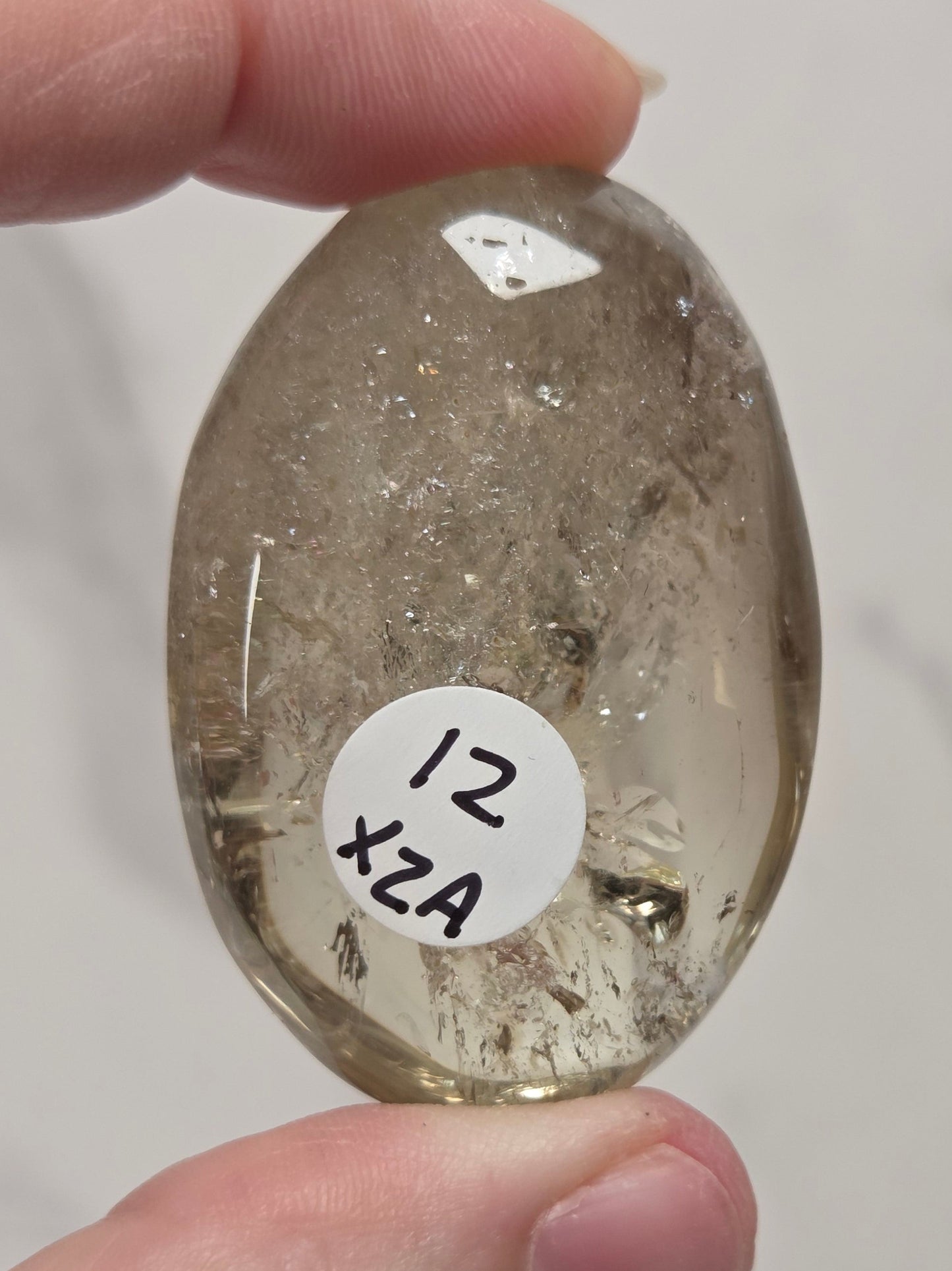 A hand holding a polished, oval-shaped smoky quartz gemstone with a white sticker labeled '12 x2A' attached to it, set against a light gray marble background.