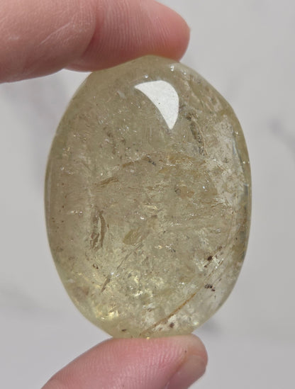 A hand holding a polished, oval-shaped clear crystal gemstone with internal inclusions against a light gray marble background.