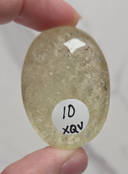 A hand holding a polished oval-shaped translucent pale yellow stone with a small white label reading '10 XQV' on the front, against a light marble-like background.
