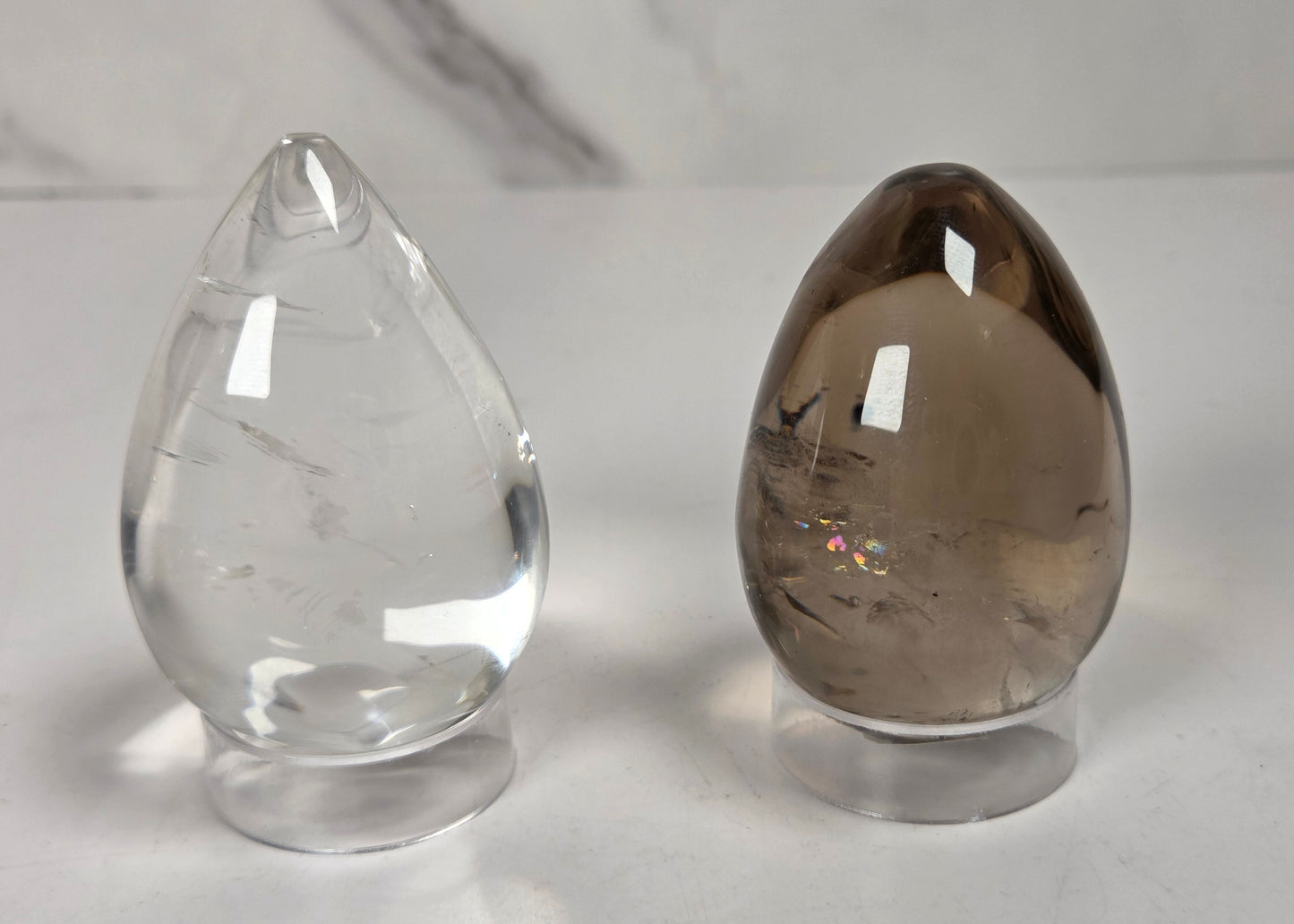 Two polished crystal ball point ornaments on clear acrylic stands, one transparent clear quartz and one smoky brown quartz, set against a white marble background.