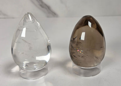 Two polished crystal ball point ornaments on clear acrylic stands, one transparent clear quartz and one smoky brown quartz, set against a white marble background.