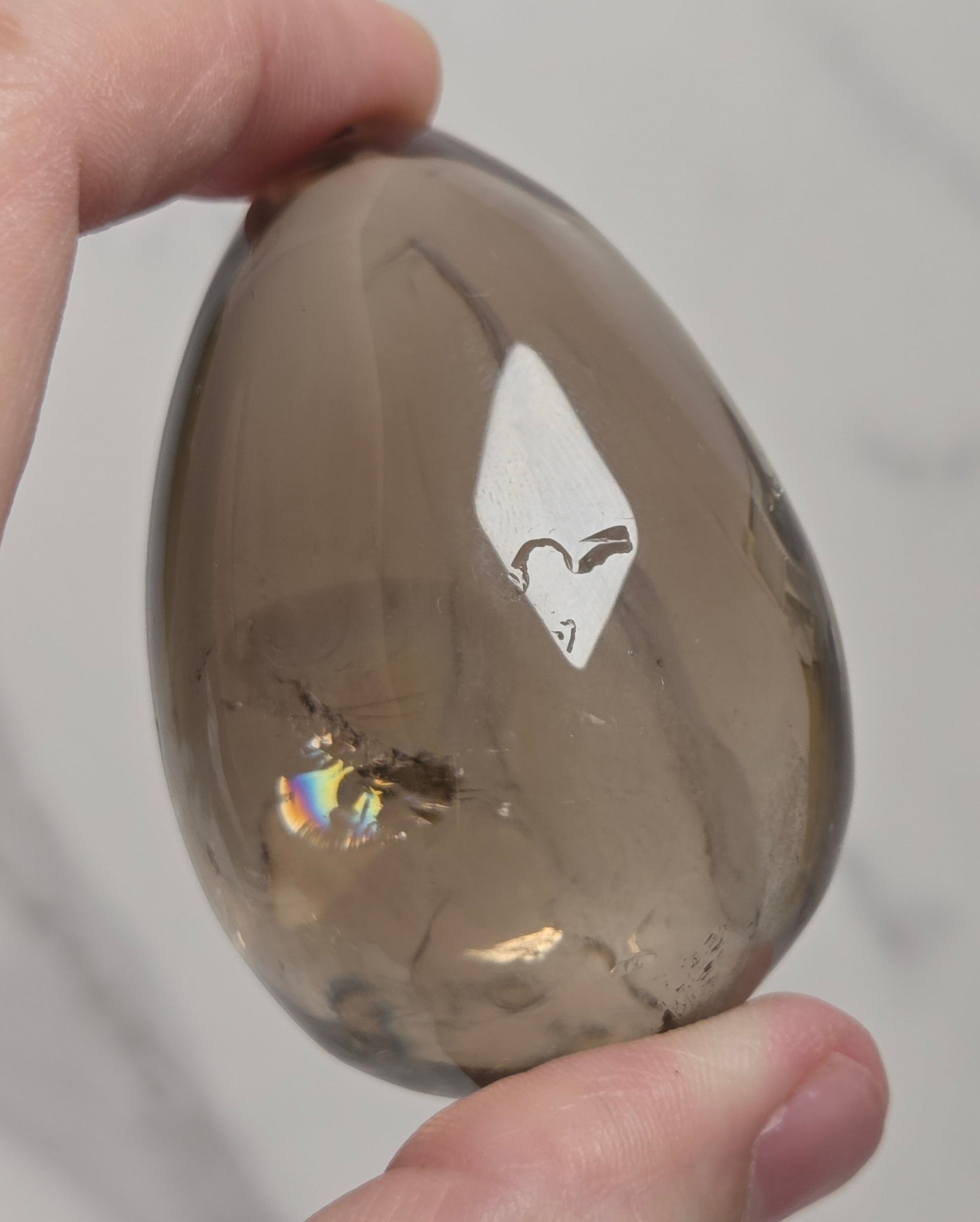 A hand holding a polished smoky quartz crystal egg against a light gray marble background.