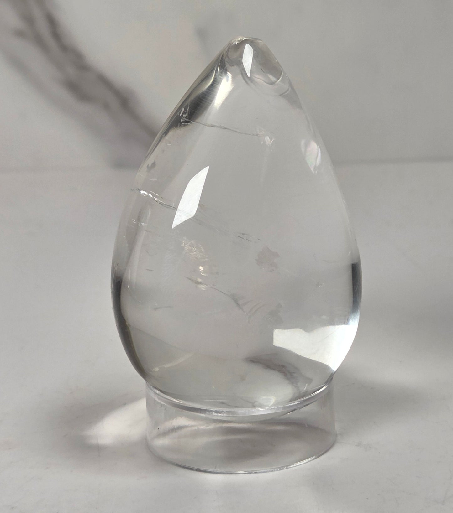 A clear teardrop-shaped glass or crystal object with a small hole near the top, resting on a small round base against a light gray background.