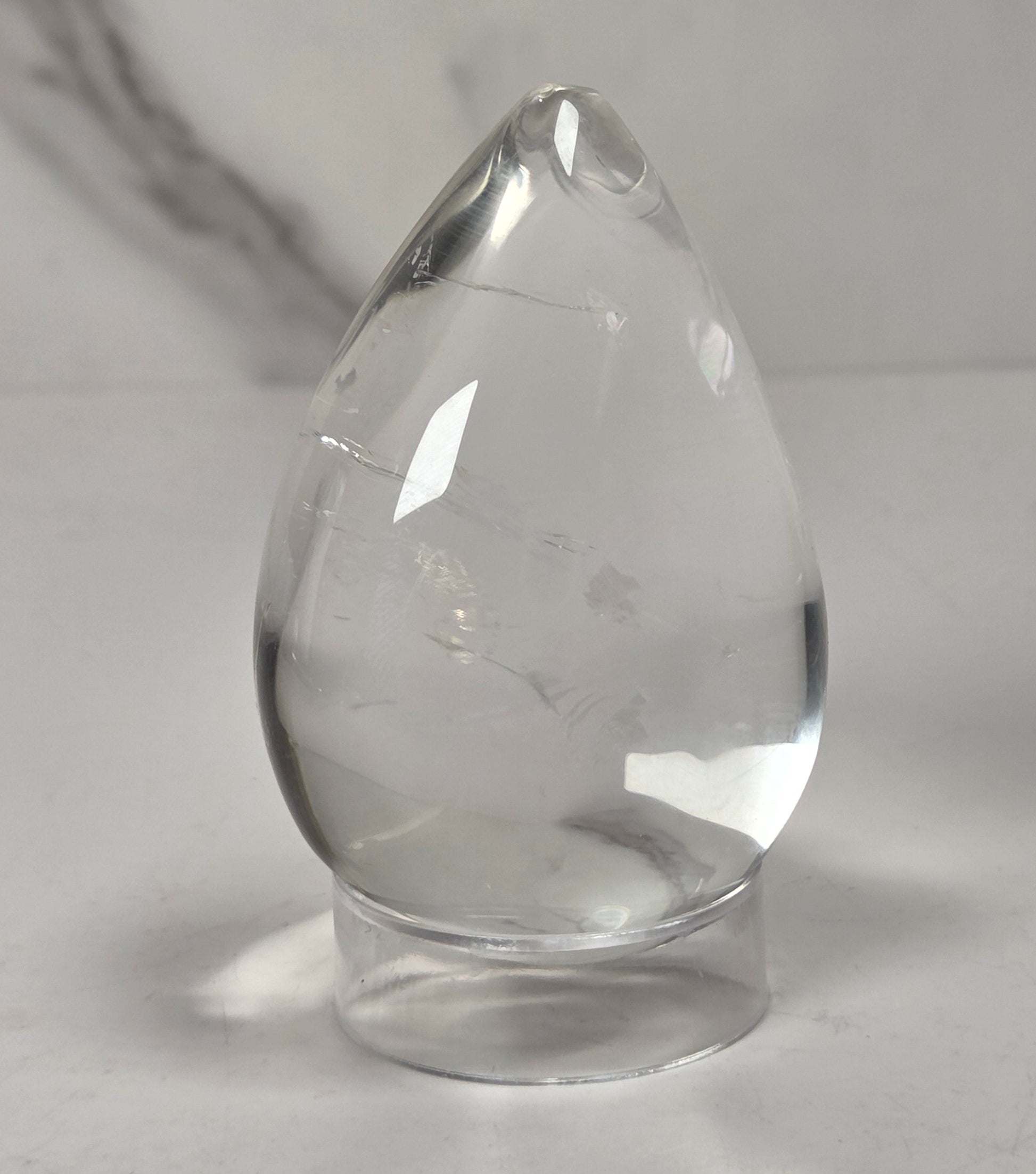A clear teardrop-shaped glass or crystal object with a small hole near the top, resting on a small round base against a light gray background.