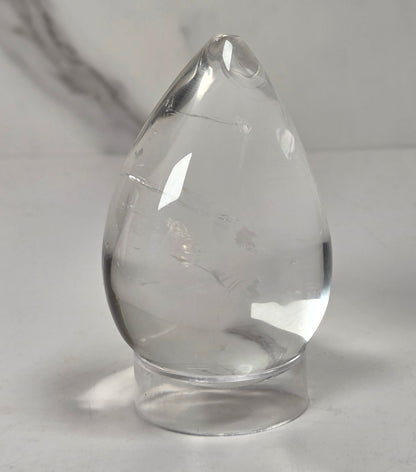 A clear teardrop-shaped glass or crystal object with a small hole near the top, resting on a small round base against a light gray background.