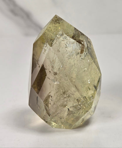 A faceted natural crystal with a pale yellow-green hue and internal inclusions, standing upright on a light gray surface.
