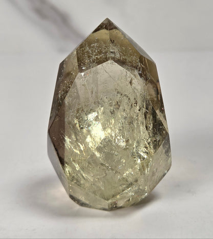 A faceted natural crystal gemstone with a pointed top, showing internal inclusions and a warm gray-brown translucent tone, photographed on a plain light background.