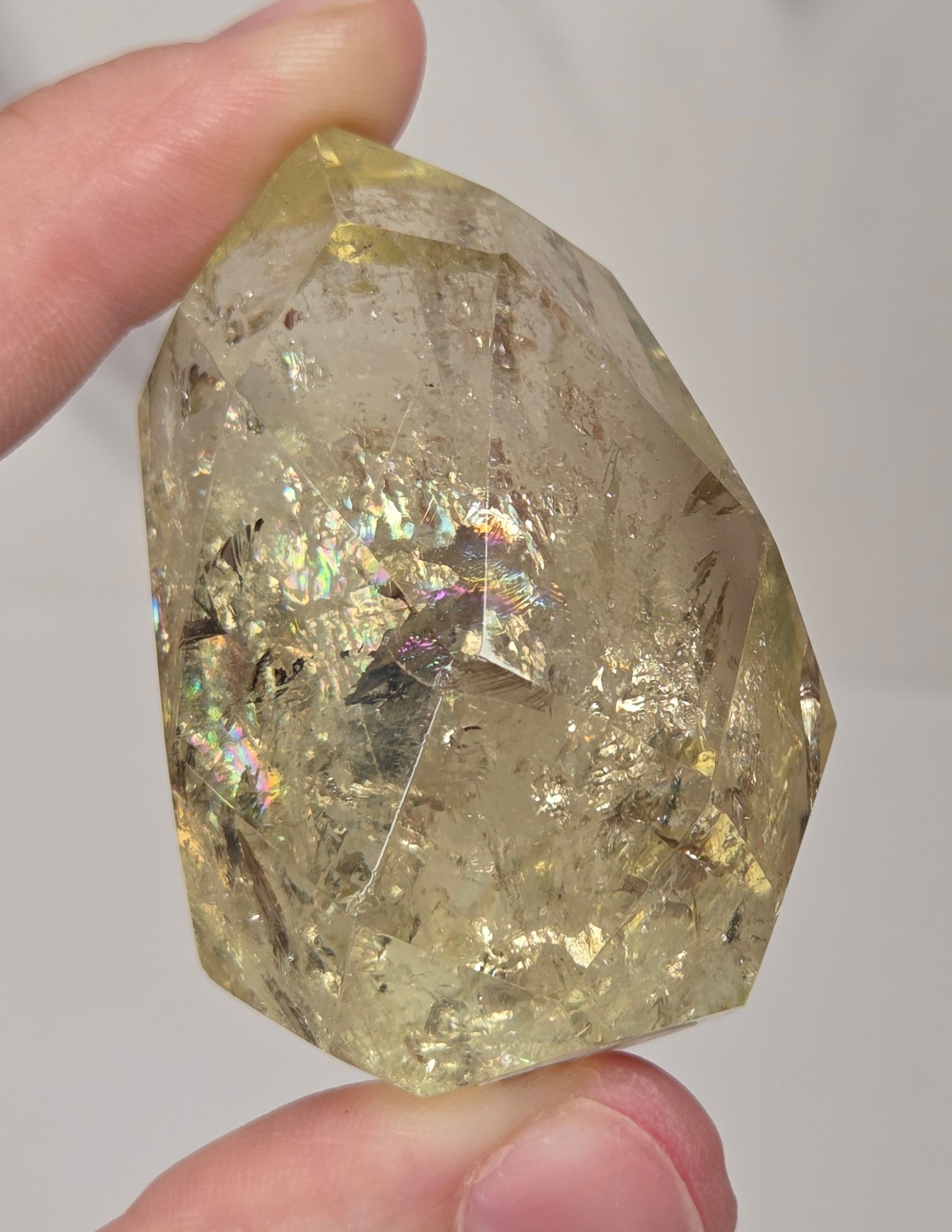 A hand holding a rough, faceted clear crystal with internal inclusions and iridescent flecks, shown against a light gray marble-like background.