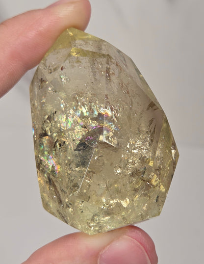 A hand holding a rough, faceted clear crystal with internal inclusions and iridescent flecks, shown against a light gray marble-like background.