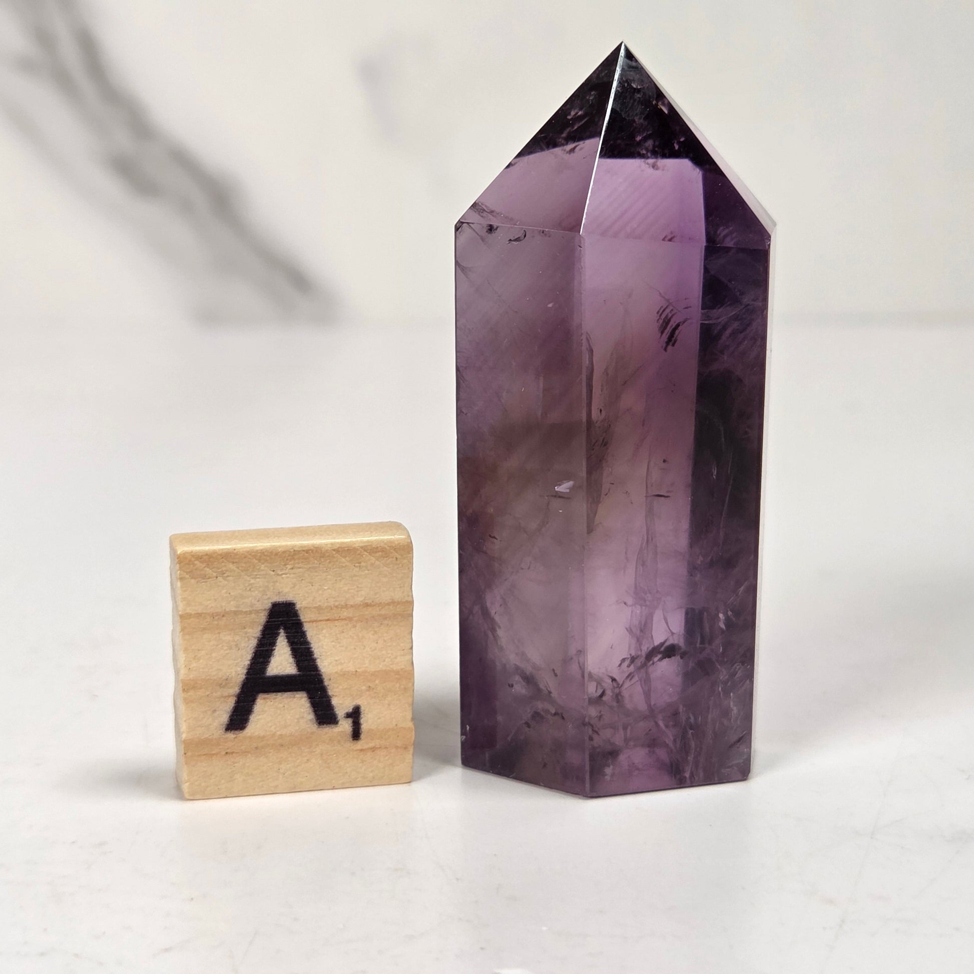 A polished purple amethyst crystal tower with a pointed top, standing next to a wooden Scrabble tile marked 'A1' on a light marble-like surface.