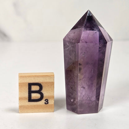 A natural purple amethyst crystal point displayed next to a wooden Scrabble tile with the letter B on a light marble surface.