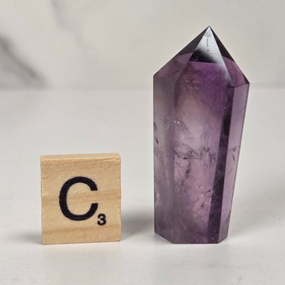 A polished purple amethyst crystal point tower standing next to a wooden Scrabble tile showing the letter 'C' and the third power, set against a light marble background.