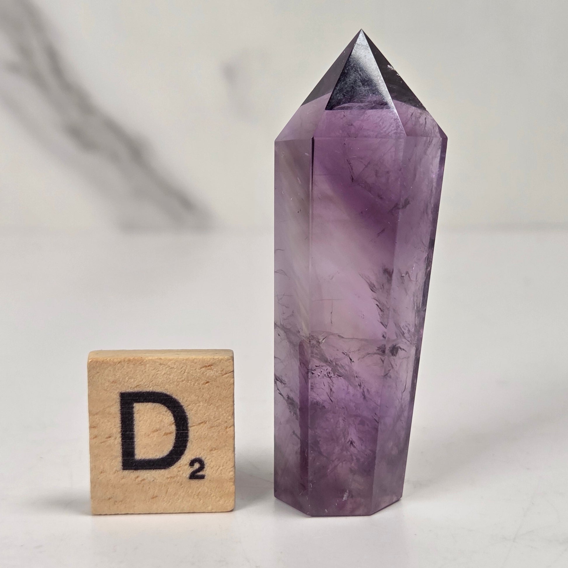 A faceted purple amethyst crystal point tower standing upright on a white surface, shown next to a wooden Scrabble tile with the letter D2.