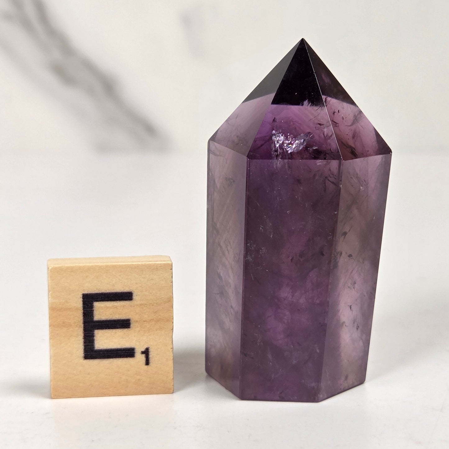 A polished purple amethyst crystal tower with a pointed top, shown next to a small wooden Scrabble tile with the letter E-1 on a light marble-like surface.