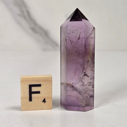 A purple amethyst crystal point tower with natural inclusions, shown next to a wooden Scrabble tile with the letter F on a light marble surface.