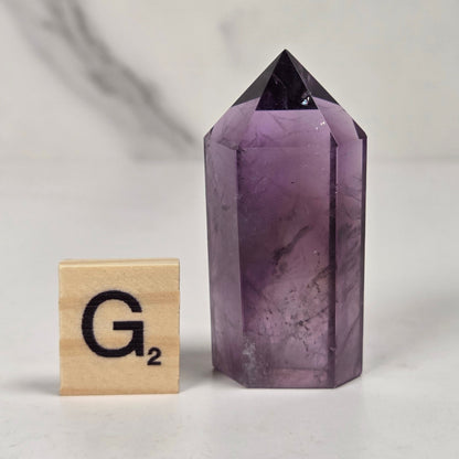 A faceted purple amethyst crystal point tower with a dark pointed top, shown next to a wooden Scrabble tile printed with 'G2' on a light marble-like surface.