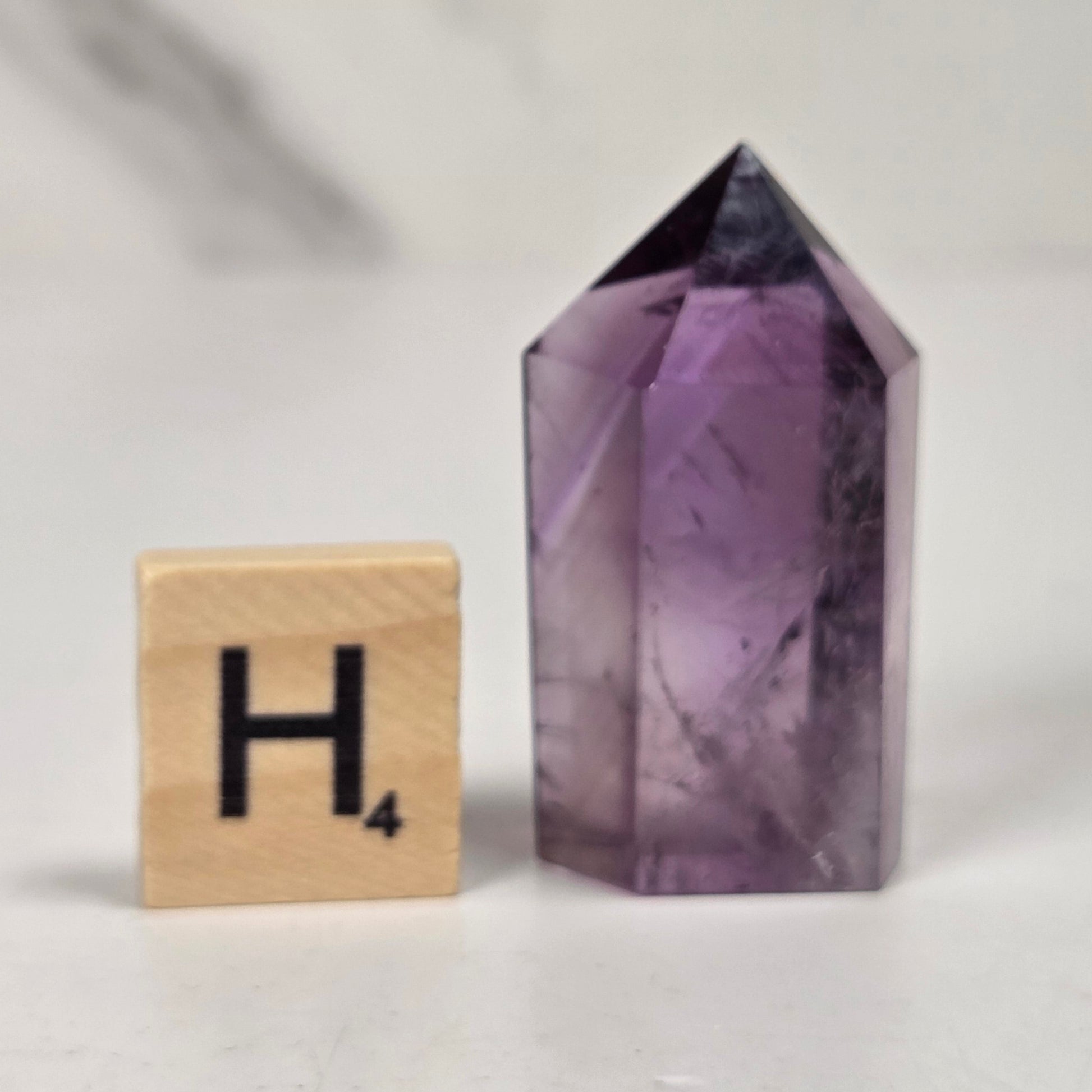 A small pointed purple crystal tower stands on a white surface next to a wooden Scrabble tile showing the letter H, with a softly blurred light gray background.