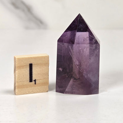 A faceted purple amethyst crystal tower with a pointed pyramid top, shown next to a small wooden block displaying the number one, on a light marble surface.