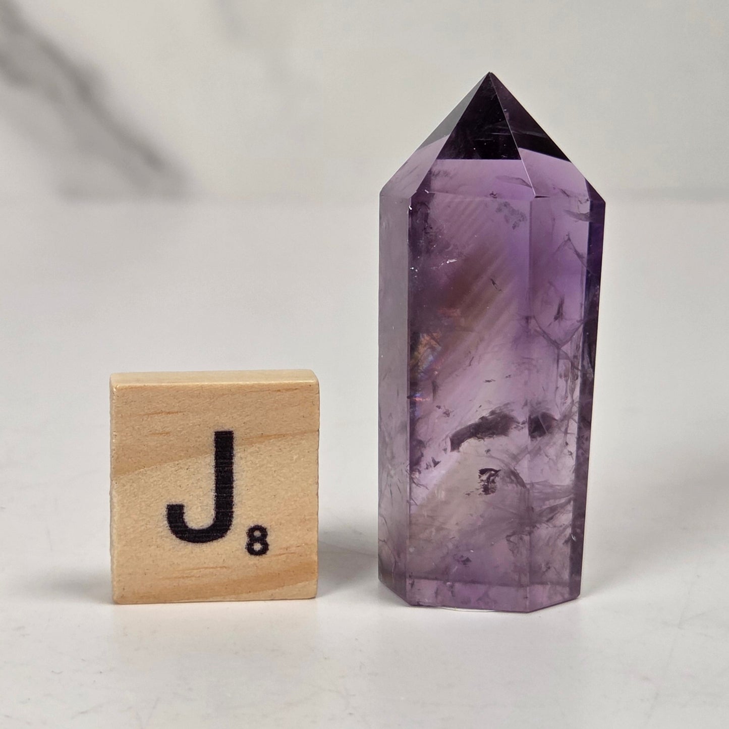 A polished purple amethyst crystal tower with a pointed top stands next to a wooden Scrabble tile showing the letter 'J' on a light gray and white marble-textured background.